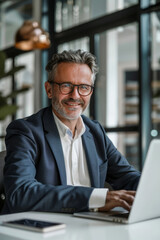 Fototapeta premium A happy businessman in a suit using a laptop at the office, sitting at a white table with glass walls in the background, wearing glasses, and looking into the camera.