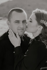 A man and woman are kissing each other. The man is wearing a ring on his finger. The woman is wearing a black dress and a headband. Scene is romantic and intimate