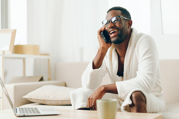 Smiling African American Freelancer Working on Laptop at Home in Bathrobe, Talking on Phone Modern...