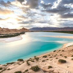 A scenic view of a turquoise blue lake surrounded by desert landscape and mountains in the background, with dramatic clouds and a setting sun in the sky