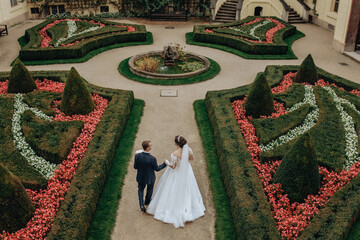 A bride and groom are walking through a garden with a fountain in the middle. The bride is wearing...