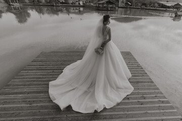 A bride is standing on a dock with a bouquet of flowers in her hand. She is wearing a white dress...