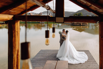 A bride and groom are standing on a dock by a lake, with the bride wearing a white dress