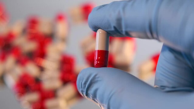 Red and white capsule in human hand in medical glove close-up on with a blurred background of a bunch of medicines pills. Healthcare and pharmacy industry concept.
