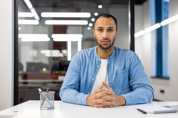 Portrait of young serious hispanic man sitting at desk in office, holding hands together and...