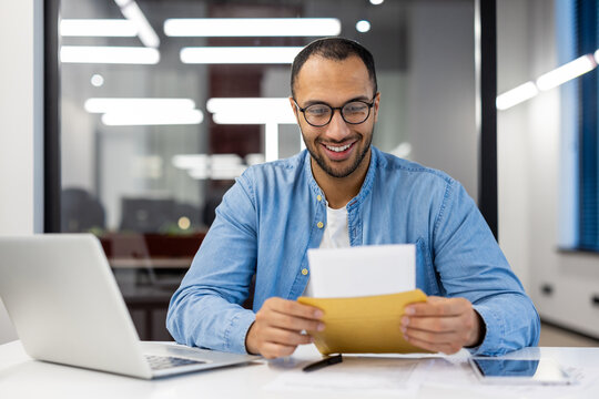 A smiling and happy young hispanic man is sitting at a desk in the office and reading an open letter in a notebook in his hands