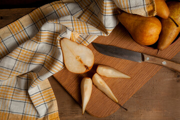 Cutting board with pears, clay bowl with fruits and yellow kitchen towel on wooden background..