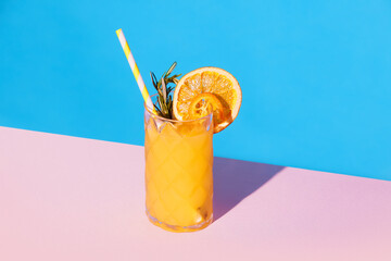 Glass with fruity lemonade with orange and rosemary, ice and straw against pink blue background....