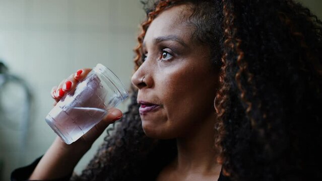 One thoughtful black middle-aged woman drinking water while gazing in the distance with pensive emotion. Contemplative latina person of African descent hydrating herself