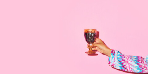 Female hand holding glass with red wine against pink background. Toast, celebration. Concept of alcohol and non-alcohol drink, party, holidays, bar, mix. Poster. Copy space for ad