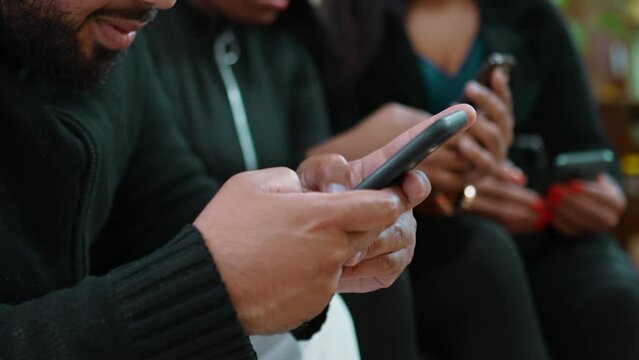 One young black latin man typing message on cellphone device with blurred friends on background. 20s person seated on couch using modern technology, holding phone adn staring at screen