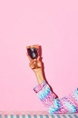 Female hand holding glass with red wine against pink background. Toast, celebration. Concept of alcohol and non-alcohol drink, party, holidays, bar, mix. Poster. Copy space for ad - obrazy, fototapety, plakaty