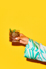 Female hand holding glass with whiskey, old-fashioned cocktail decorated with mint against bright yellow background. Concept of alcohol drink, party, holidays, bar, mix. Poster. Copy space for ad - obrazy, fototapety, plakaty