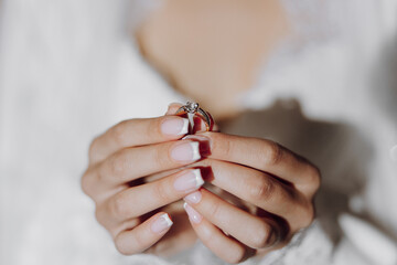 A woman is holding a ring in her hand. The woman is wearing a white dress and has her nails...