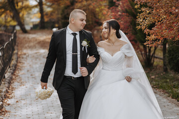A bride and groom are walking down a path in a park. The bride is wearing a white dress and the...