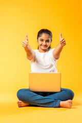 portrait of Indian asian schoolgirl girl using laptop, sitting cross-legged isolated on yellow background