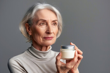 Portrait of an elderly pretty American woman with a jar of cosmetic cream in her hand on gray background. Concept of cosmetics for adult people. Silver economy concept.