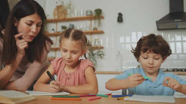 Mother attentively helps her two young children with their homework at a kitchen table, nurturing role of parents in children's education