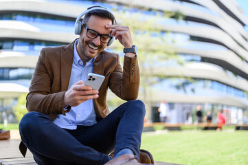 Cheerful young businessman in headphones watching video over mobile phone against building in city