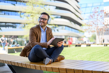 Smiling professional with headphones and digital tablet looking away and sitting on bench in city