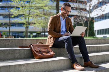 Smiling male entrepreneur checking e-mails over laptop while sitting with bag on steps in city