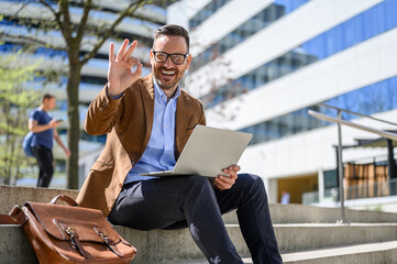 Portrait of positive male professional showing OK sign while working over laptop on steps in city
