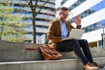 Happy businessman waving hand while attending online meeting over laptop while sitting on steps