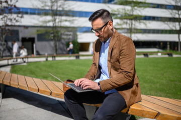 Serious entrepreneur in glasses working on project over laptop while sitting on bench in modern city
