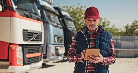 Handsome mature Caucasian man wearing red cap standing next to trucks while using tablet device....