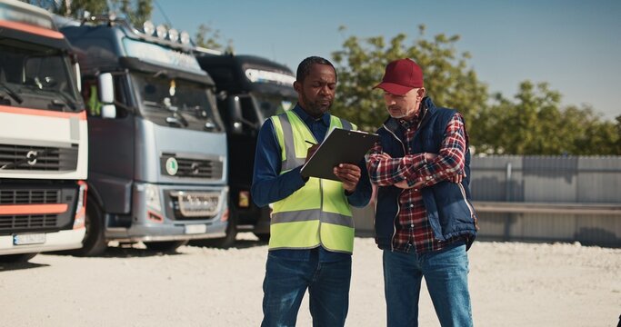 Hardworking mixed-race people discussing about upcoming import goods on their trucks. Busy manager with vest writing on clipboard paper some information about departure of trucks for delivery abroad.