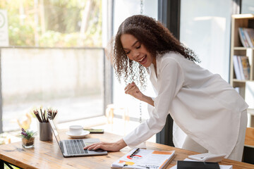 A woman is sitting at a desk with a laptop and a cup of coffee. She is smiling and she is happy