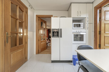 Frontal image of a kitchen with white furniture with metal handles and a column with a refrigerator...