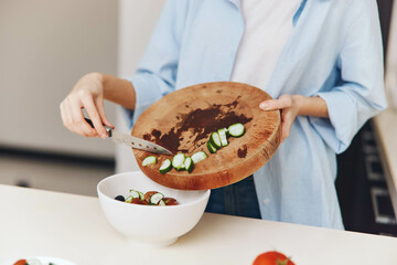 Woman preparing fresh salad with cucumbers and tomatoes on a wooden cutting board in blue shirt