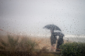 portrait of people walking on border the sea with umbrella behind a window with rain drops