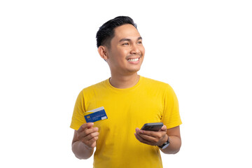 Happy young Asian man holding mobile phone and bank credit card, looking aside with big smile isolated on white background