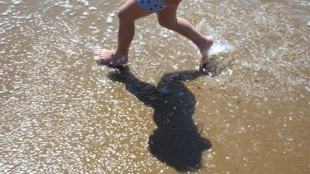 Close-up of a three-year-old boy's feet running along the beach and splashing in the waves in slow motion.