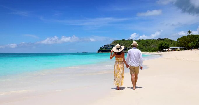 Happy holiday couple with sunhats walks down a tropical beach with turquoise sea in the Caribbean, Antigua island