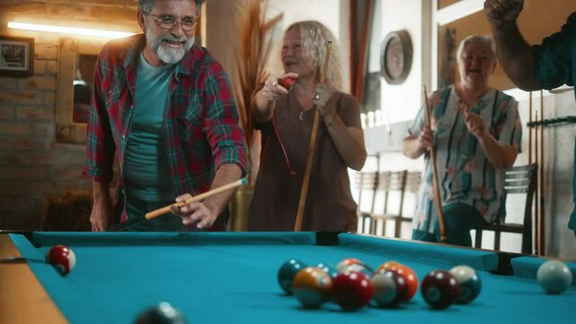 Elderly 60s man play Billiard with friends, break pyramid of balls on a Billiard Pool table. Group of seniors playing Billiards home