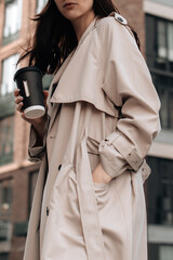 Young stylish woman wearing beige long seasonal trench coat walking around the city with a cup of coffee. Street style cloth concept
