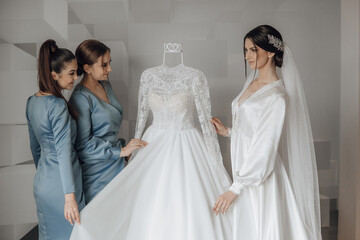 Naklejka premium Three women are standing in front of a white wedding dress. One of the women is holding the dress up to the other two. Scene is one of excitement and anticipation for the upcoming wedding