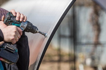 A man holds a battery-powered screwdriver and assembles a new greenhouse made of polyethylene,...