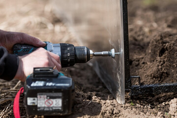 A man holds a battery-powered screwdriver and assembles a new greenhouse made of polyethylene, polycarbonate. Macro photography. Side view. Preparing for the spring garden season.