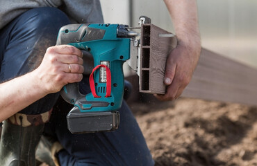 A man holds a battery-powered electric saw and builds new beds in a greenhouse made of wood-polymer...