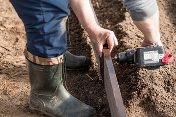 A man holds a battery-powered screwdriver and collects new beds from WPC into a greenhouse made of polyethylene, polycarbonate. Macro photography. Side view. Preparing for the spring garden season.