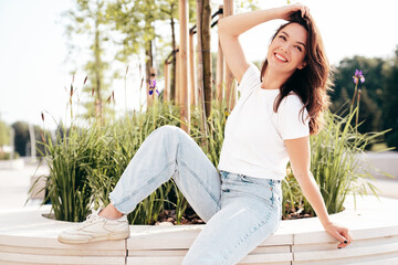 Young beautiful smiling hipster woman in trendy summer white t-shirt and jeans clothes. Sexy carefree model posing on the street background at sunset. Positive model outdoors