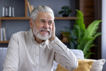 Close-up portrait of an older man suffering from severe ear pain, sitting on the sofa at home and...