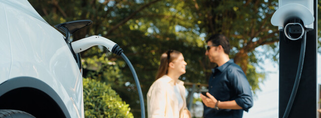 Young couple recharge electric car battery from charging station in green city park in springtime....
