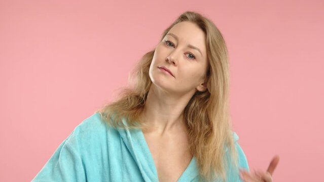 This photo shows a young woman in a light blue robe, carefully combing her long blonde hair, set against a soft pink background, highlighting a daily beauty routine. 
