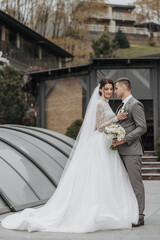Fototapeta premium A bride and groom are posing for a picture in front of a building. The bride is wearing a white dress and the groom is wearing a gray suit. They are holding a bouquet and a vase