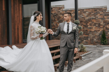 A bride and groom are standing outside of a building, with the bride holding a bouquet and the groom wearing a tie